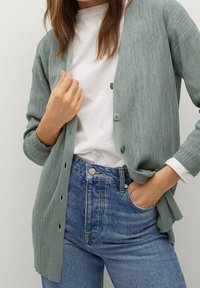 Cardigan verde chiaro a costine con bottoni, indossato sopra una camicia bianca. Abbinato a jeans blu a vita alta. Texture morbida, vestibilità rilassata.