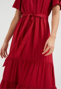 Červené šaty s krátkým rukávem, nařaseným pasem a detailem mašle. Mají vrstvenou sukni s texturovanou tkaninou a jemným vzorem.