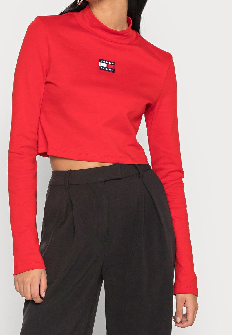 Czerwony krótki t-shirt z długim rękawem i stójką oraz małym prostokątnym haftowanym logo w kolorach niebieskim i białym, noszony na czarnych spodniach z wysokim stanem.