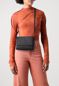 Borsa a tracolla nera con finitura testurizzata, chiusura a patta e tracolla regolabile, indossata sopra una maglietta a maniche lunghe color ruggine e pantaloni rosa chiaro.