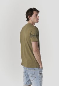 Maglietta verde oliva con un motivo grafico sulla spalla, abbinata a pantaloncini di jeans azzurri. Il tessuto sembra morbido e casual.