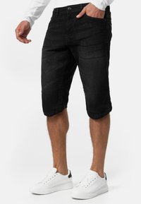 Crne traperice kratke hlače s opuštenim krojem, s preklopljenim rubovima i dizajnom s pet džepova. Nosene s bijelim tenisicama, na bijeloj pozadini.