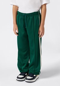 Ein Kind trägt grüne Jogginghosen mit weißen Seitenstreifen, ein weißes T-Shirt und schwarz-weiße Sneakers und steht vor einem weißen Hintergrund.