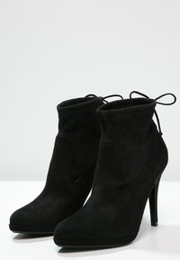 Botines de ante negro con tacón alto, que presentan un suave puño y un diseño de punta afilada. Detalle de lazo ajustable en la parte trasera.