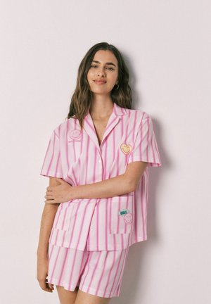 Femme aux longs cheveux foncés portant un t-shirt à manches courtes rayé rose et blanc et un short assorti avec des patchs visage souriant, debout devant un fond clair.