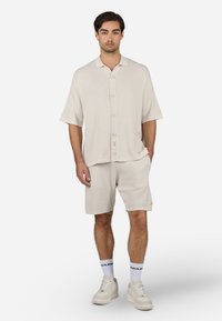 Camisa de manga corta de canalé beige con botones en el frente, combinada con pantalones cortos a juego. Lleva zapatos deportivos blancos y calcetines con texto negro.
