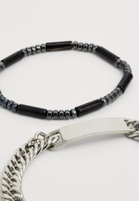 Pulsera de cuentas negras con acentos metálicos cilíndricos, acompañada de una pulsera de cadena de plata que presenta una placa de metal rectangular y lisa.