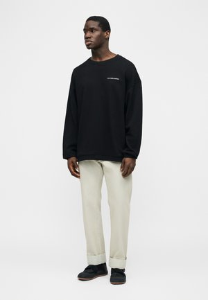 Νεαρός άνδρας στέκεται, φοράει μαύρο πουλόβερ με μακριά μανίκια και το κείμενο "LES BENJAMINS" και ανοιχτόχρωμο μπεζ παντελόνι με μανσέτες και μαύρα παπούτσια.