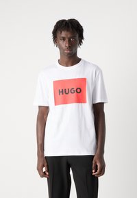 Hvit t-skjorte med korte ermer som har en rød rektangulær logo med ordet "HUGO" i fet svart tekst i midten. Laget av bomull.