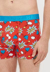 Sarkanas bokserbikses ar zilu elastīgu jostiņu, uz kuras rakstīts "HAPPY." Dizainā ir komiksu stila grafikas ar sirdīm, mākoņiem un izlaušanos.