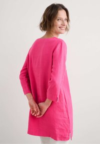 Fuchsia linnen tuniek met driekwart mouwen, zijsplitten en een losse pasvorm. De stof heeft een zachte textuur en een lichte, ademende kwaliteit.