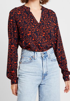 Femme portant un chemisier bleu marine avec un motif floral rouge, rentré dans un jean taille haute bleu clair sur un fond uni.