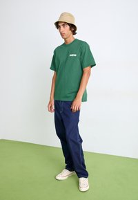 Grünes Baumwoll-T-Shirt mit "Dickies"-Logo, blaue Jeans, beiger Bucket Hat und weiße Turnschuhe auf einem grünen Boden vor einem hellen Hintergrund.