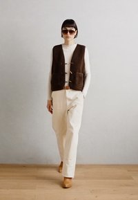 Brown wool vest with two front pockets, worn over a white ribbed top. Cream-colored trousers and brown shoes. Model stands against a plain wall.