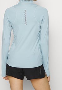 Hellblaues Langarmoberteil mit hohem Kragen, das ein Zickzackmuster auf dem Rücken und Netzpaneelen an den Seiten aufweist, kombiniert mit schwarzen Shorts.