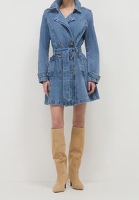 Denim trench coat in light blue with double-breasted buttons, waist tie, and slanted pockets; paired with knee-high suede beige boots.