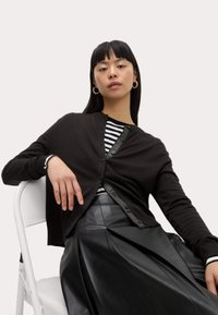 Cardigan noir avec des boutons, porté sur un haut à rayures noir et blanc, associé à une jupe plissée noire. Modèle assis sur une chaise blanche.