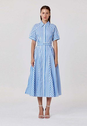 Femme portant une robe midi bleue et blanche à rayures verticales, boutonnée, à manches courtes et ceinture, associée à des sandales blanches à talons hauts.