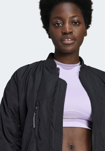 Fekete bomber dzseki cipzáros elől, bordázott gallérral és oldalsó zsebekkel. Egy világos lila sportfelső felett viselve, amelyen egy diszkrét logó találhat�ó.