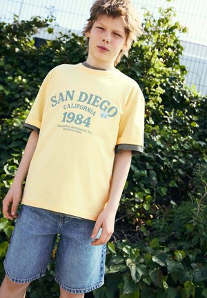 Junge trägt gelbes, geschichtetes T-Shirt mit dem Aufdruck „San Diego California 1984“ und Jeansshorts, steht draußen vor grünem Laub.