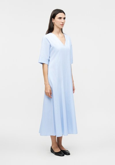 Γυναίκα που στέκεται με ανοιχτό γαλάζιο φόρεμα μέχρι το γόνατο, με μανίκια μέχρι τους αγκώνες και μαύρα επίπεδα παπούτσια, μπροστά σε απλό λευκό φόντο.