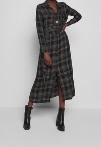 Abito camicia lungo a scacchi in tonalità scure, con una chiusura a bottoni frontale, due tasche sul petto, una vita definita da una cintura e spacchi laterali. Indossato con stivaletti neri.
