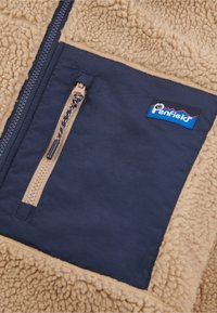 Tan fleece jacket with navy nylon accents, featuring a zippered pocket, textured material, and a logo patch.