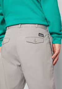 Šedé kalhoty s texturovanou tkaninou, které mají dvě zadní kapsy s knoflíkovými uzávěry a cedulkou na pravé kapse. Z výše je vidět zelený svetr.