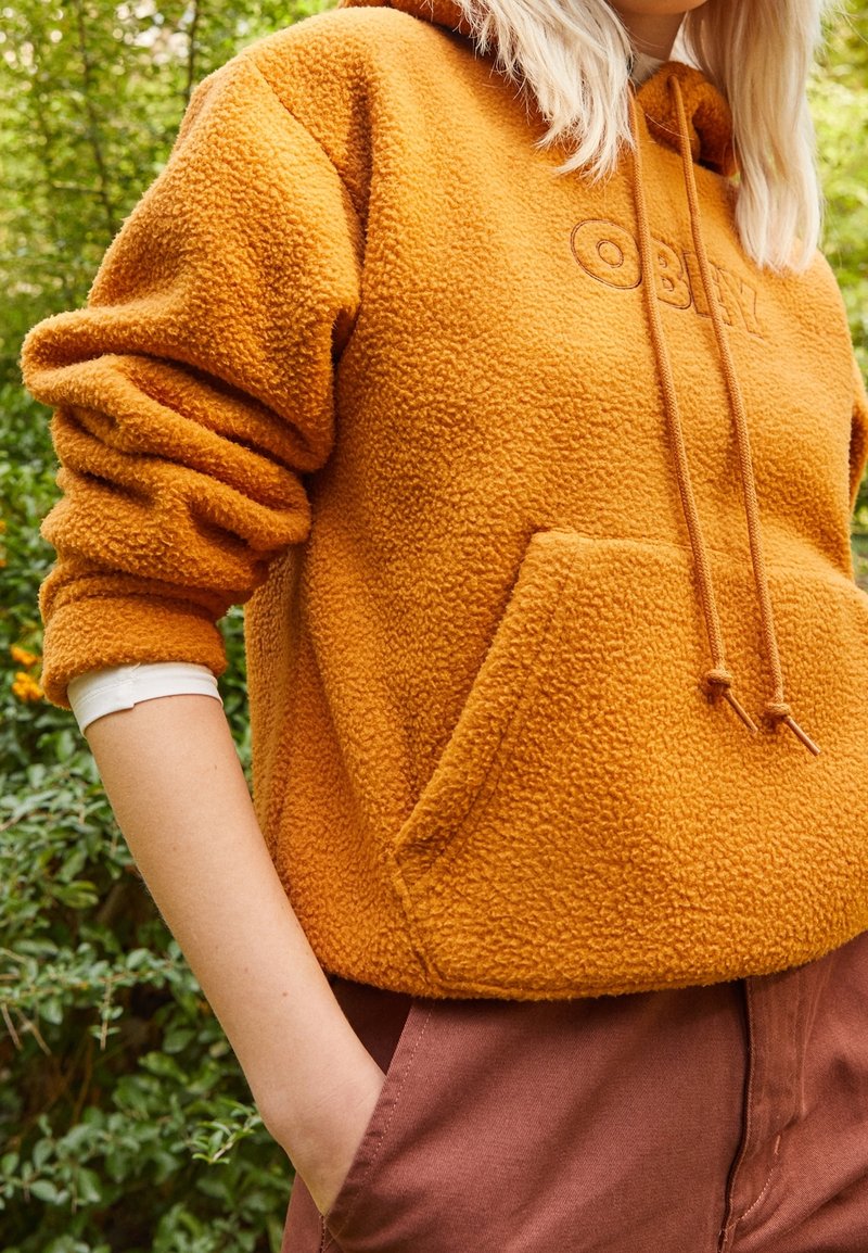 Fleece-pullover hoodie i senapsgul med en frampå ficka, snörning och präglad logotyp. Bärs över bruna byxor. Grön växtlighet i bakgrunden.