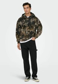 Haut à capuche à motif camouflage dans des tons terreux avec un cordon de serrage, associé à un pantalon noir et des baskets en toile. Le hoodie a une coupe décontractée et des poches latérales.