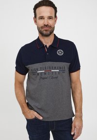 Paita polo dengan kerah biru navy dan lengan pendek, badan abu-abu dengan teks yang dicetak, serta logo bulat di dada sebelah kiri.
