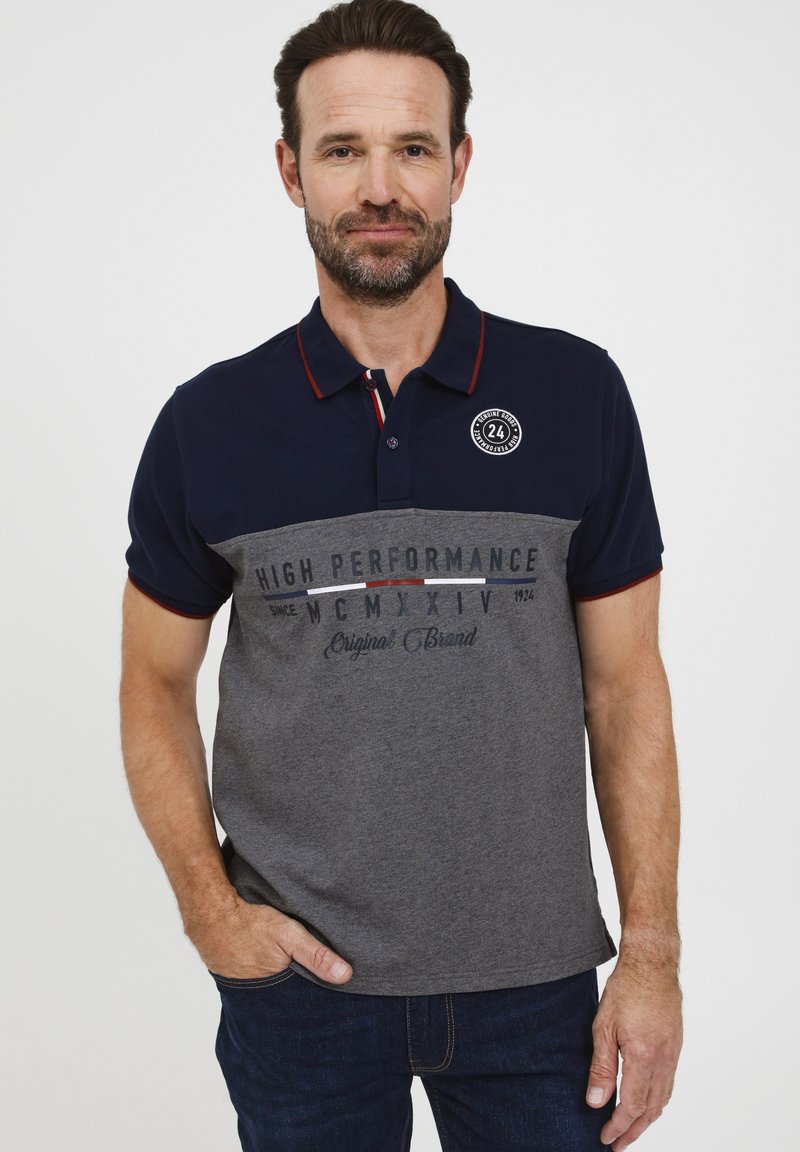 Paita polo dengan kerah biru navy dan lengan pendek, badan abu-abu dengan teks yang dicetak, serta logo bulat di dada sebelah kiri.