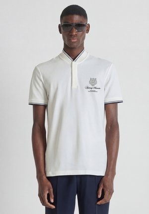 Hombre con gafas de sol negras, camisa polo blanca de manga corta con pequeño logo en el pecho y pantalones oscuros, de pie contra un fondo liso.