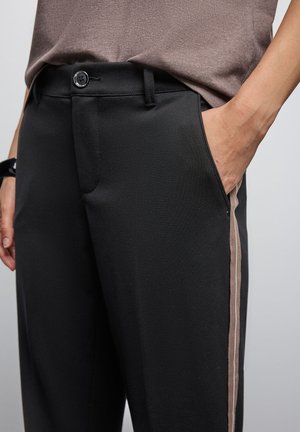 Schwarze Hose mit glatter Textur, ausgestattet mit einem seitlichen Streifen in Beiǵe. Hat eine Fronttasche und einen Knopfverschluss an der Taille.