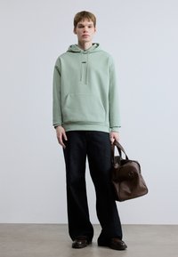 Ανοιχτοπράσινο φούτερ με τσέπη μπροστά, συνδυασμένο με σκούρο τζιν φαρδιάς γραμμής. Καφέ δερμάτινη τσάντα με στρογγυλεμένο σχήμα, κρατημένη στο δεξί χέρι.