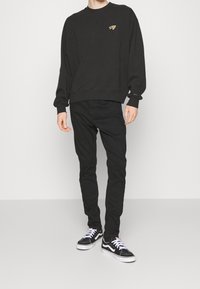 Musta huppari pyöreällä kauluksella ja kultaisella logolla, yhdistetty mustiin slim fit -farkkuihin ja mustiin lenkkareihin, joissa on valkoiset nauhat ja yksityiskohdat.