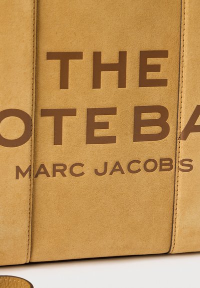 Ταμπά σουέντ επιφάνεια με μεγάλο καφέ κείμενο που διαβάζεται μερικώς ως «THE NOTE BA» και μικρότερο κείμενο «MARC JACOBS» από κάτω.