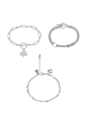 Trois bracelets en chaîne argentée, l'un avec un fermoir à bascule et un design multi-brins, un autre avec un charm en forme de trèfle, et un dernier avec de petits charms en forme de cœur et de cloche.