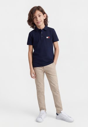 Chłopiec z włosami do ramion, ubrany w granatową koszulkę polo, beżowe spodnie i białe tenisówki, stojący z jedną ręką w kieszeni.