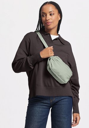 Frau trägt einen dunkelbraunen Pullover mit Kragen und blaue Jeans, trägt eine hellgrüne Umhängetasche über der Schulter.