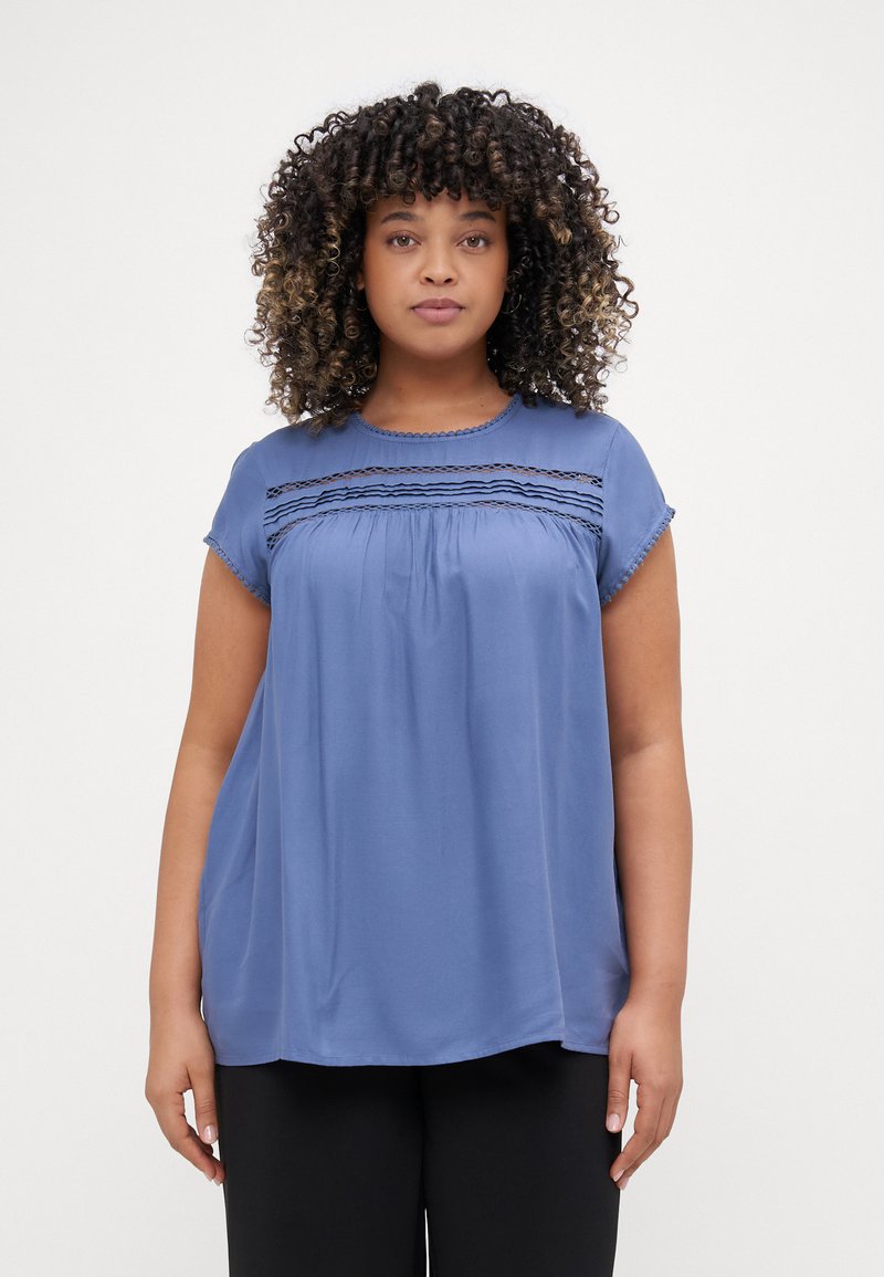 Blue short-sleeve blouse with embroidered detail near the neckline, flowing silhouette, and smooth fabric texture. Paired with black pants.