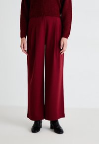Burgundy wide-leg trousers made from soft fabric, featuring front pleats and a smooth texture, paired with a fitted black shoe.