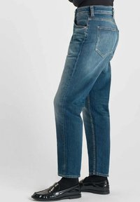 Vaqueros de mezclilla azul oscuro con un corte recto, diseño de cinco bolsillos y una cinturilla clásica, combinados con zapatos negros brillantes y calcetines.