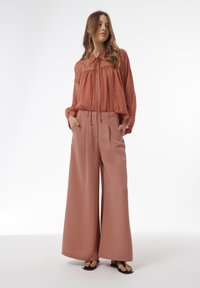 Roze, loszittend blouse met een gerimpelde halslijn en brede, hoge taille broek in een bijpassende kleur, gecombineerd met zwarte flip-flops.