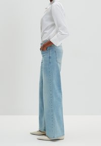 Osoba ubrana w jasnoniebieskie spodnie z szerokimi nogawkami, białe buty na szpilce o szpiczastym nosku oraz białą koszulę z długim rękawem zapinaną na guziki, stoi z rękami w kieszeniach.