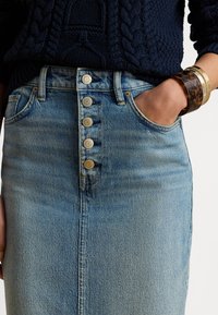 Γυναίκα που φοράει μπλε πουλόβερ πλεκτό με πλεξούδες και μπλε τζιν φούστα με ψηλή μέση και πέντε μεταλλικά κουμπιά, με το χέρι στην τσέπη, φορώντας βραχιόλια από χελώνα και χρυσό.