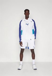 Hvit treningsdrakt med jakke som kan glidelåses opp, med blå detaljer og logo, kombinert med matchende shorts, fullført med hvite joggesko.