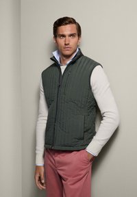 Grünes Steppweste mit Reißverschluss, hohem Kragen und kontrastierenden Schulterdetails. Getragen über einem weißen Pullover mit blauen Akzenten.