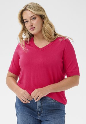 Blonde Frau trägt ein knallpinker V-Ausschnitt Kurzarmshirt und blaue Jeans, steht mit verschränkten Händen auf der Taille vor einem schlichten Hintergrund.