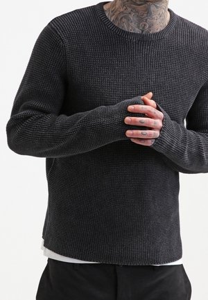 Man die een donkergrijze, gestructureerde gebreide trui met duimgaten draagt, handen samenvouwt, met zichtbare tatoeages op de nek en handen.